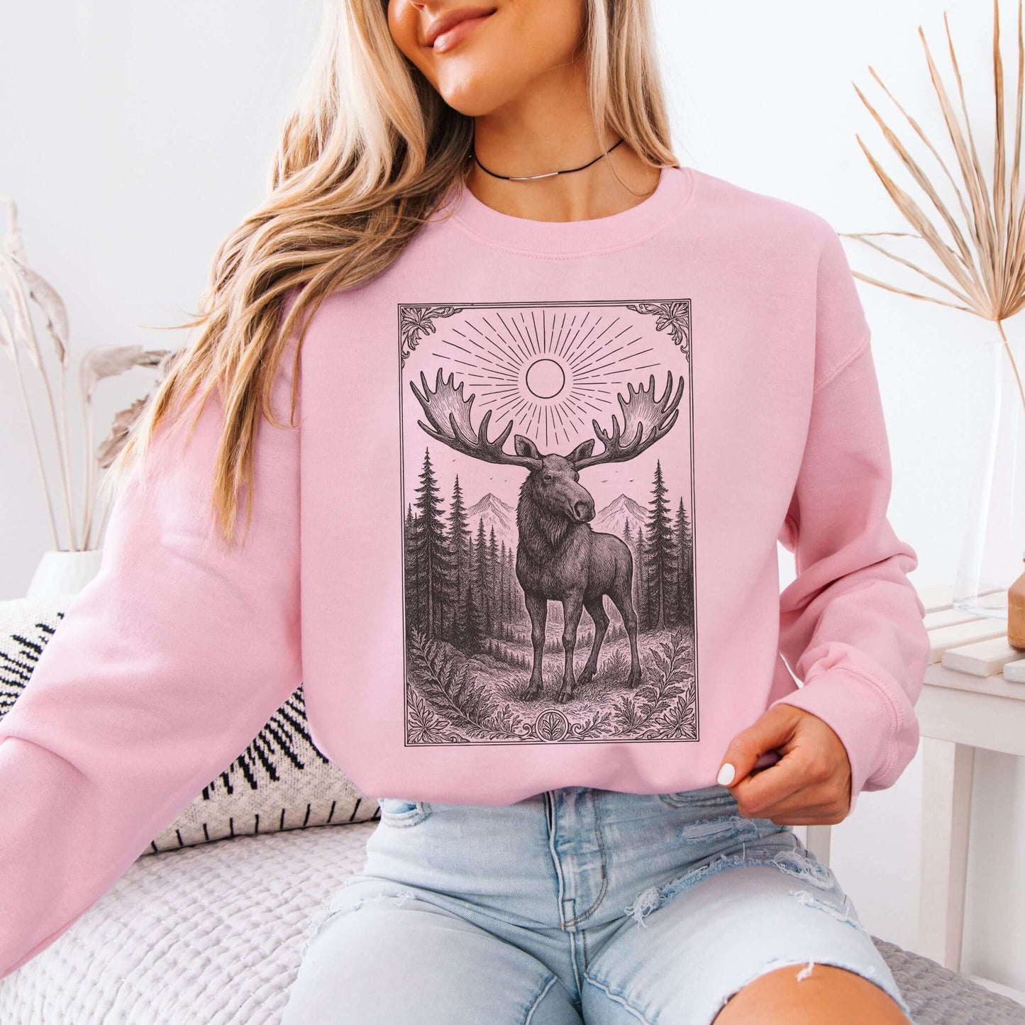 Model wearing pink Mystic Moose crewneck sweatshirt featuring a moose graphic in a forest setting.