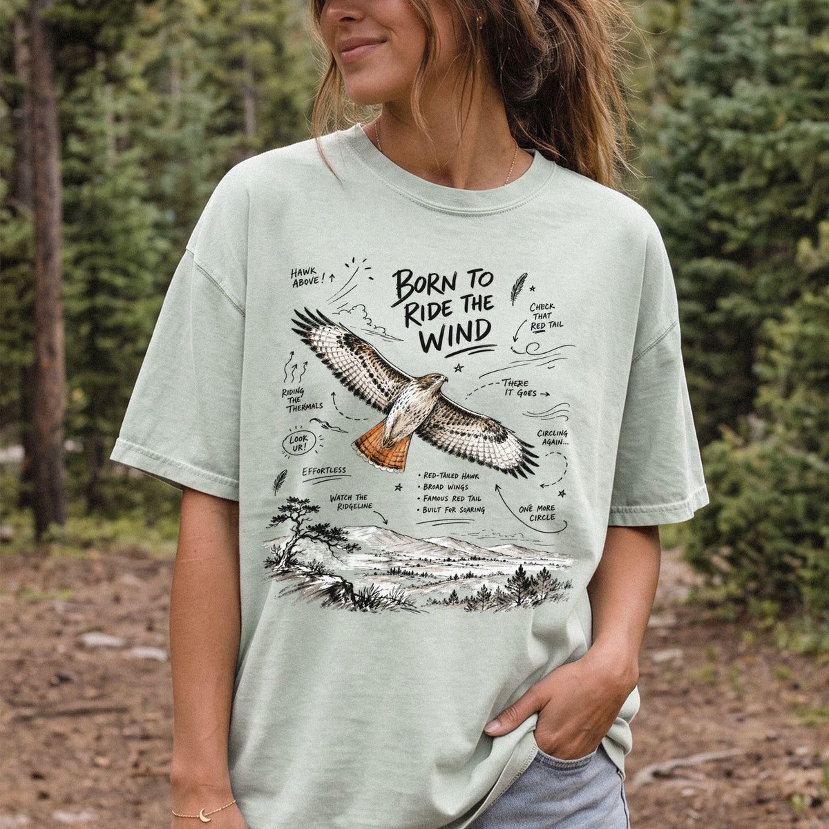 Born to Ride the Wind Comfort Colors Shirt – Red-Tailed Hawk design of a Red-Tailed Hawk illustrated in flight above an open ridgeline, with field-note style callouts and a scenic landscape below, printed on a Bay T-shirt, worn by a woman.