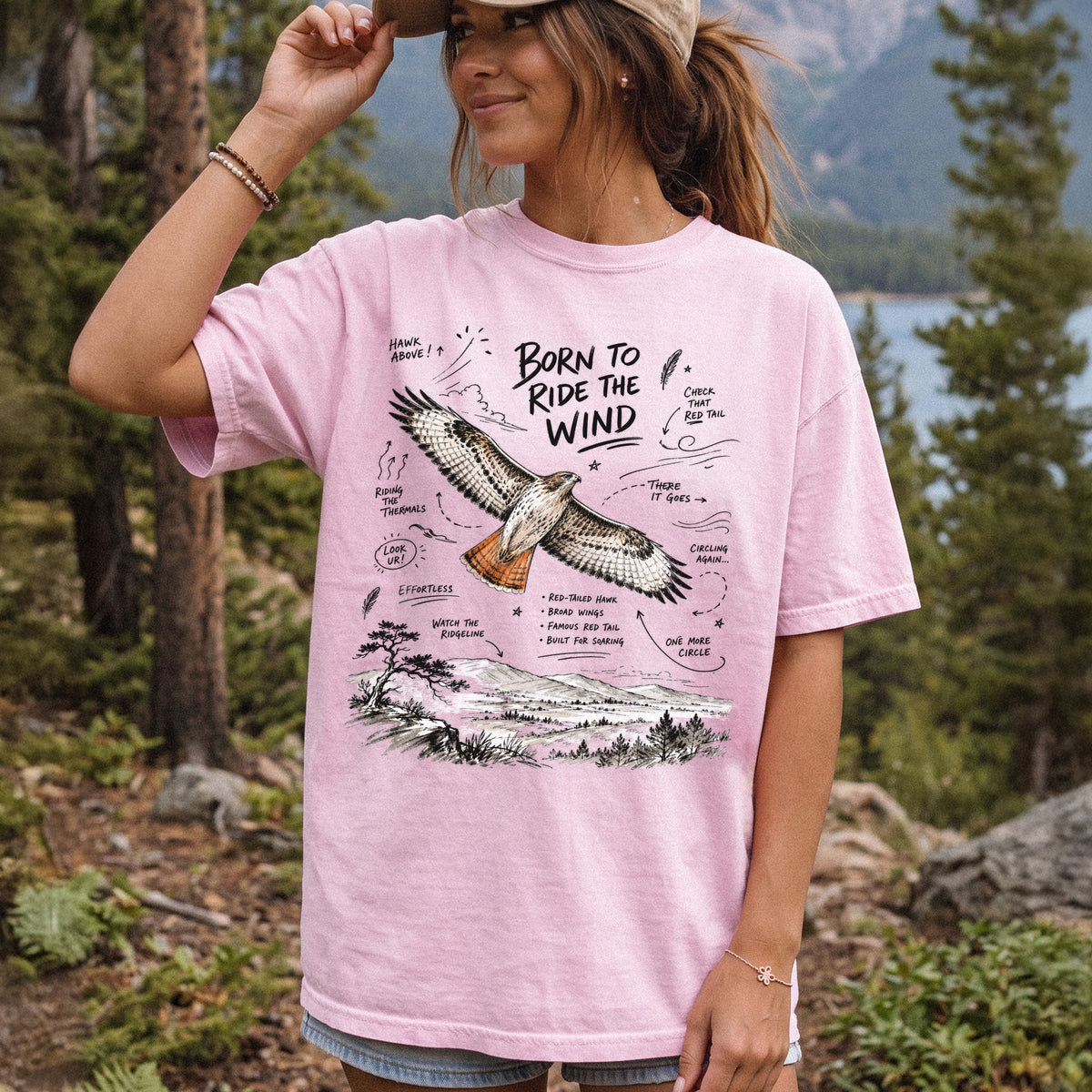 Born to Ride the Wind Comfort Colors Shirt – Red-Tailed Hawk design of a Red-Tailed Hawk illustrated in flight above an open ridgeline, with field-note style callouts and a scenic landscape below, printed on a Blossom T-shirt, worn by a woman.
