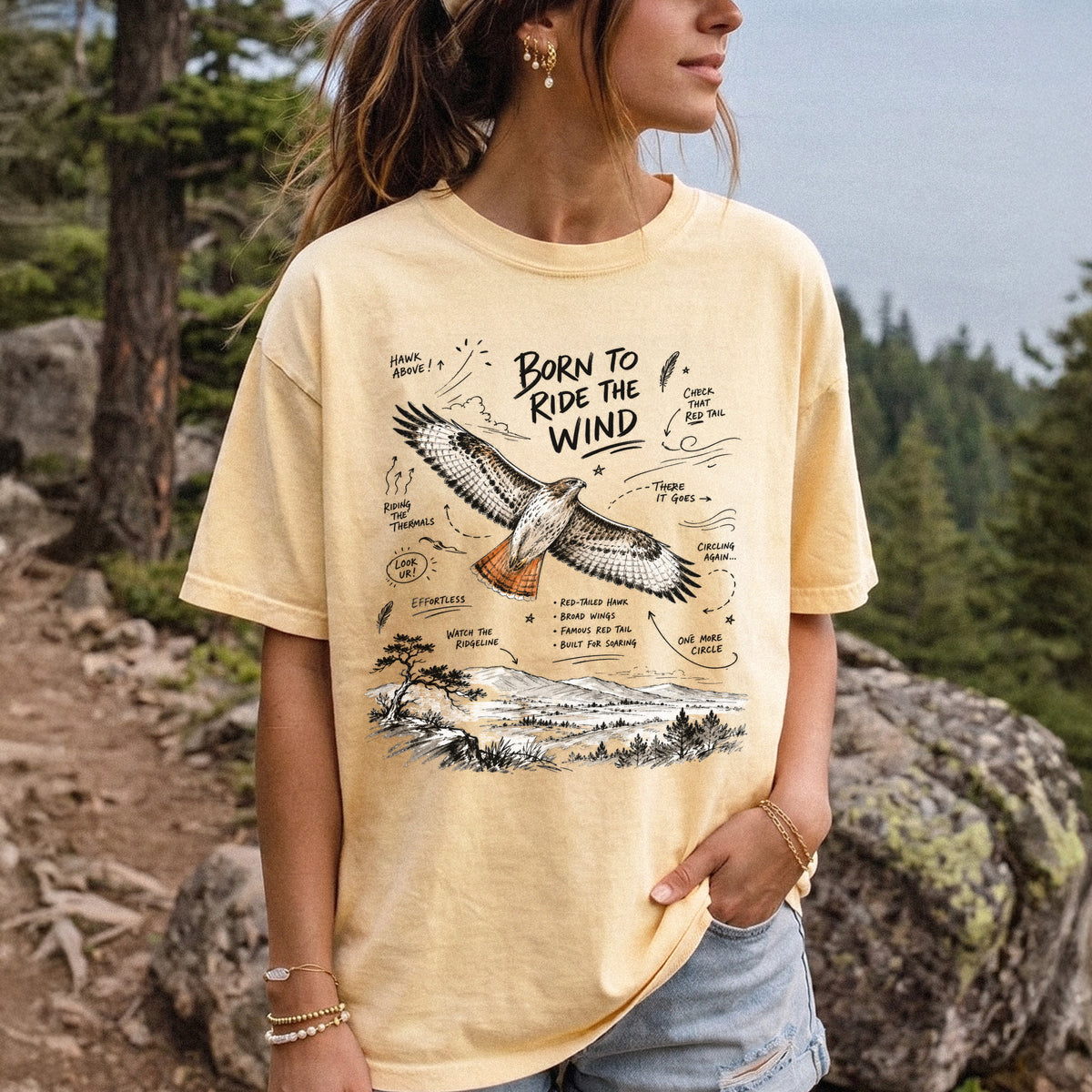 Born to Ride the Wind Comfort Colors Shirt – Red-Tailed Hawk design of a Red-Tailed Hawk illustrated in flight above an open ridgeline, with field-note style callouts and a scenic landscape below, printed on a Butter T-shirt, worn by a woman.