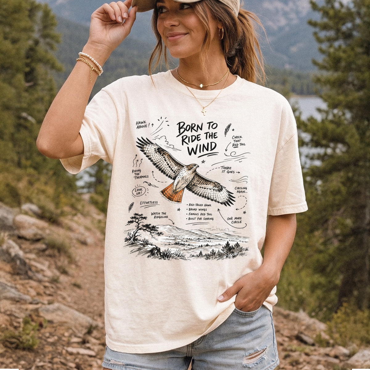 Born to Ride the Wind Comfort Colors Shirt – Red-Tailed Hawk design of a Red-Tailed Hawk illustrated in flight above an open ridgeline, with field-note style callouts and a scenic landscape below, printed on an Ivory T-shirt, worn by a woman.