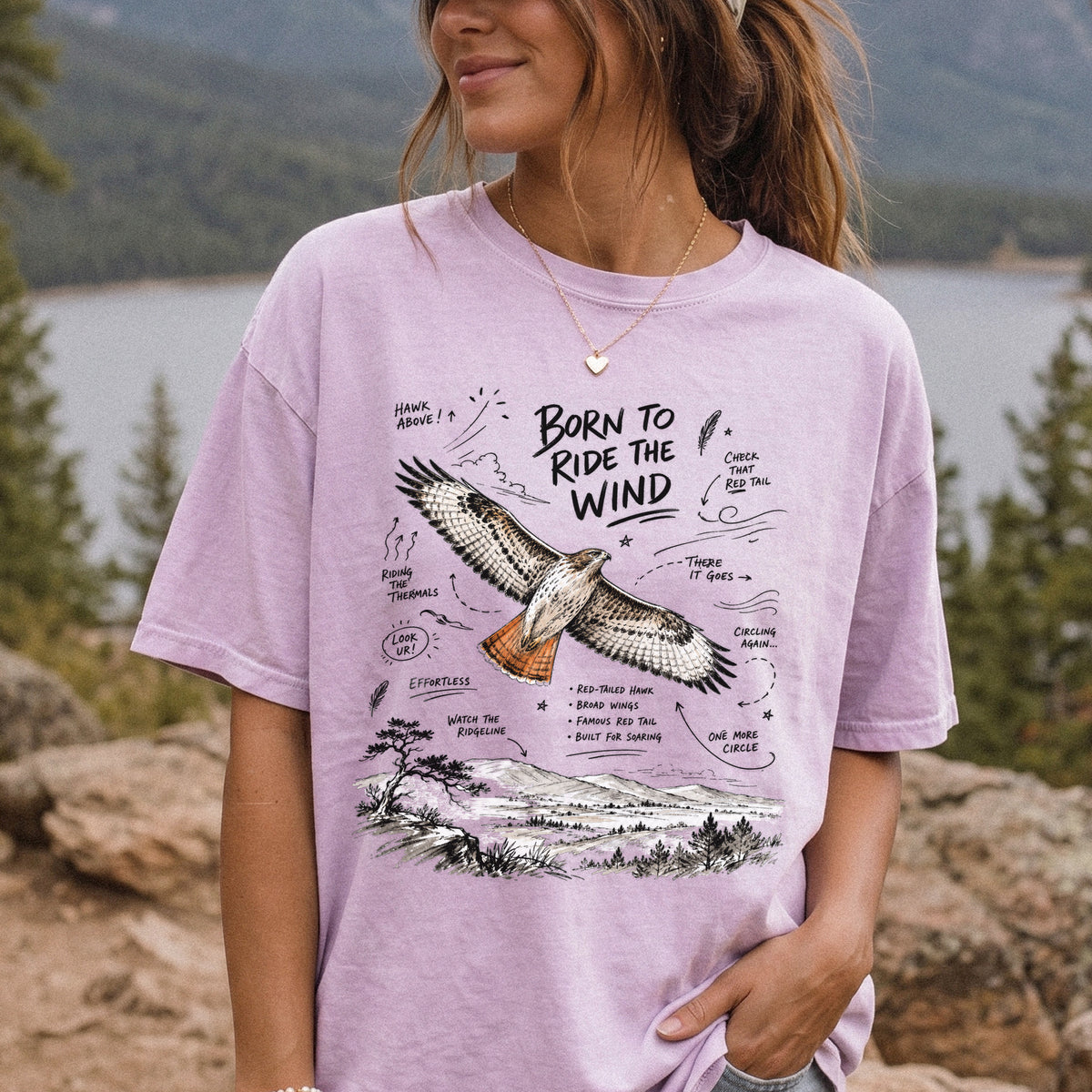 Born to Ride the Wind Comfort Colors Shirt – Red-Tailed Hawk design of a Red-Tailed Hawk illustrated in flight above an open ridgeline, with field-note style callouts and a scenic landscape below, printed on an Orchid T-shirt, worn by a woman.