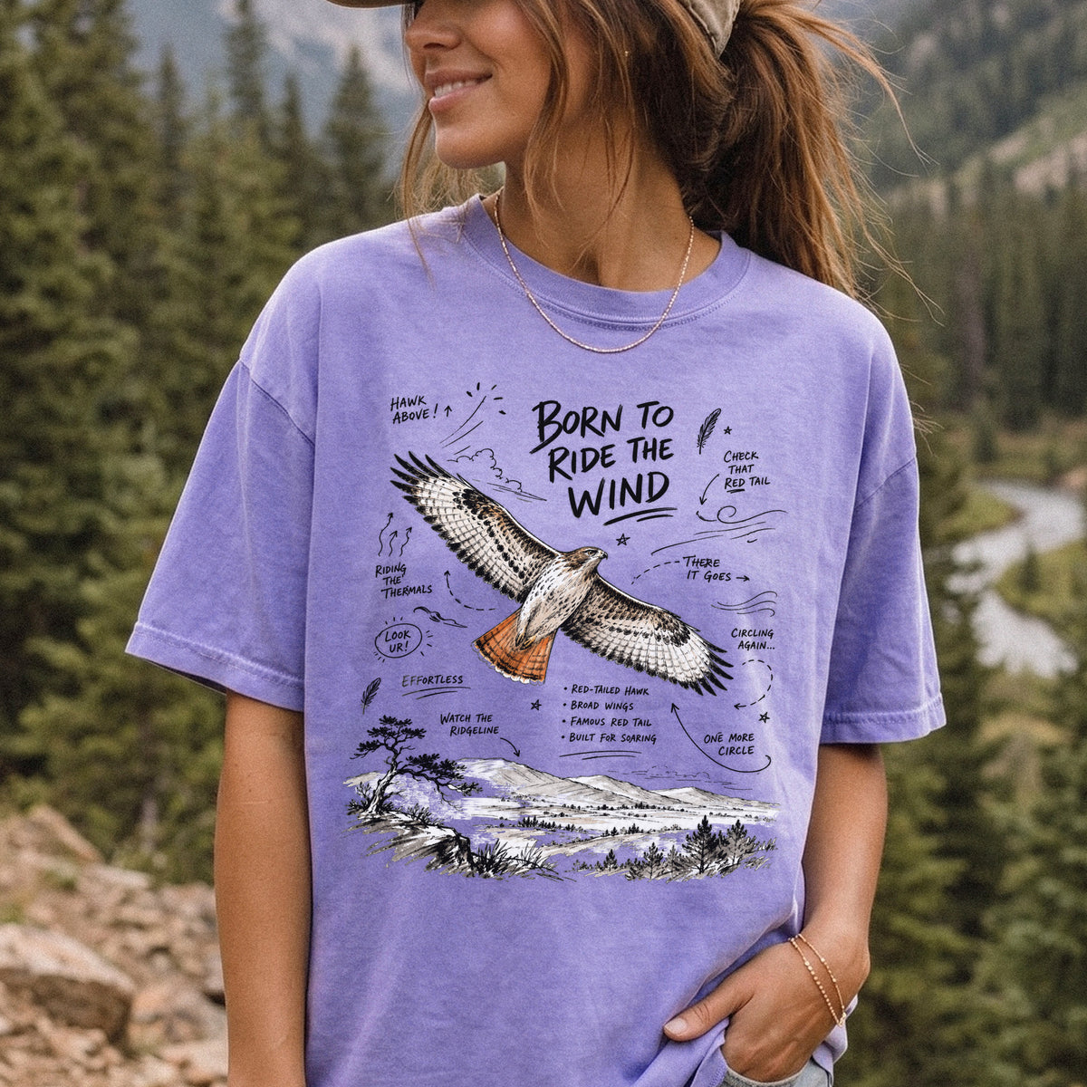 Born to Ride the Wind Comfort Colors Shirt – Red-Tailed Hawk design of a Red-Tailed Hawk illustrated in flight above an open ridgeline, with field-note style callouts and a scenic landscape below, printed on a Violet T-shirt, worn by a woman.