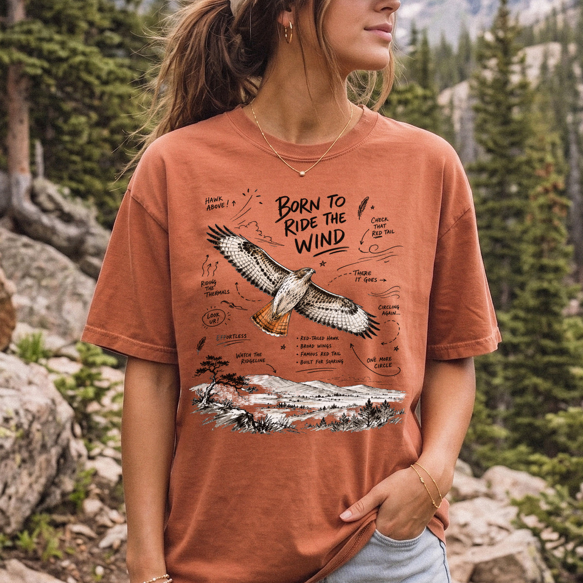 Born to Ride the Wind Comfort Colors Shirt – Red-Tailed Hawk design of a Red-Tailed Hawk illustrated in flight above an open ridgeline, with field-note style callouts and a scenic landscape below, printed on a Yam T-shirt, worn by a woman.