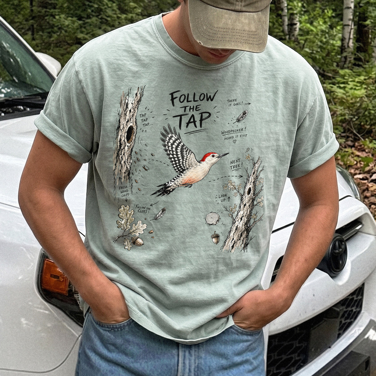 Follow the Tap Comfort Colors Shirt – Red-Bellied Woodpecker design of a Red-Bellied Woodpecker illustrated in flight, surrounded by tree trunks, wood chips, feathers, oak leaves, and acorn details, printed on a Bay T-shirt.