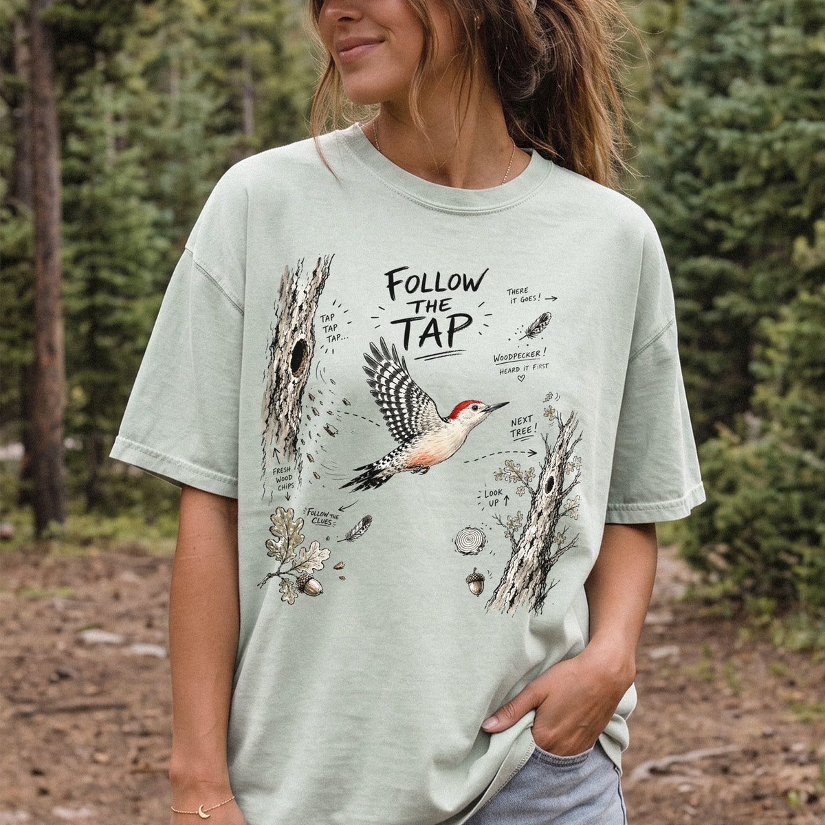 Follow the Tap Comfort Colors Shirt – Red-Bellied Woodpecker design of a Red-Bellied Woodpecker illustrated in flight, surrounded by tree trunks, wood chips, feathers, oak leaves, and acorn details, printed on a Bay T-shirt, worn by a woman.