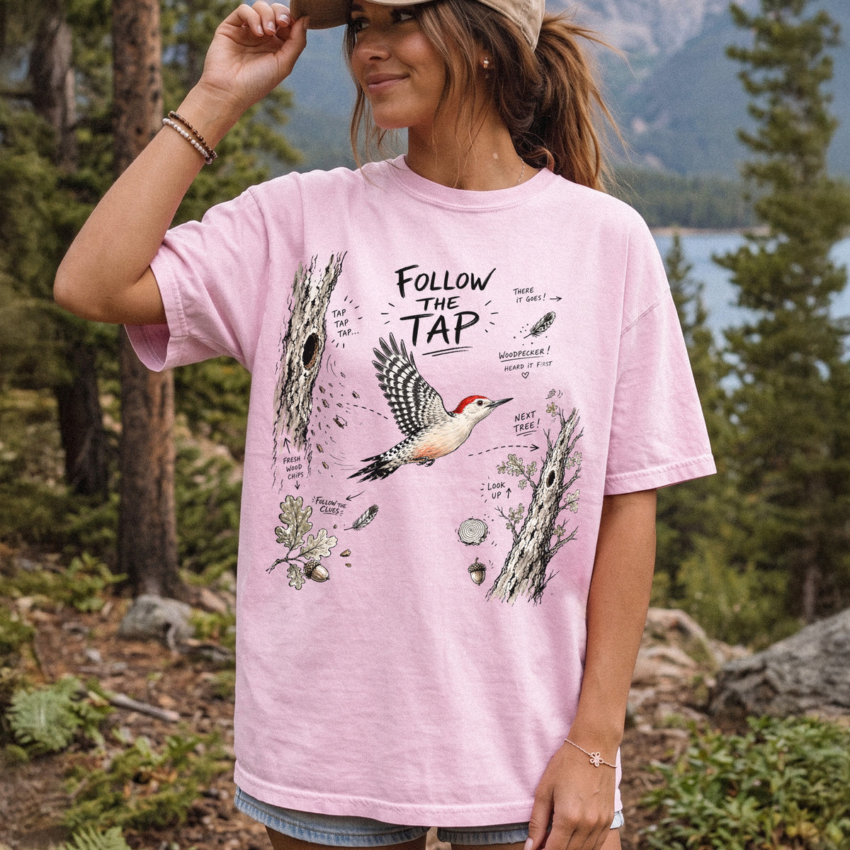 Follow the Tap Comfort Colors Shirt – Red-Bellied Woodpecker design of a Red-Bellied Woodpecker illustrated in flight, surrounded by tree trunks, wood chips, feathers, oak leaves, and acorn details, printed on a Blossom T-shirt, worn by a woman.