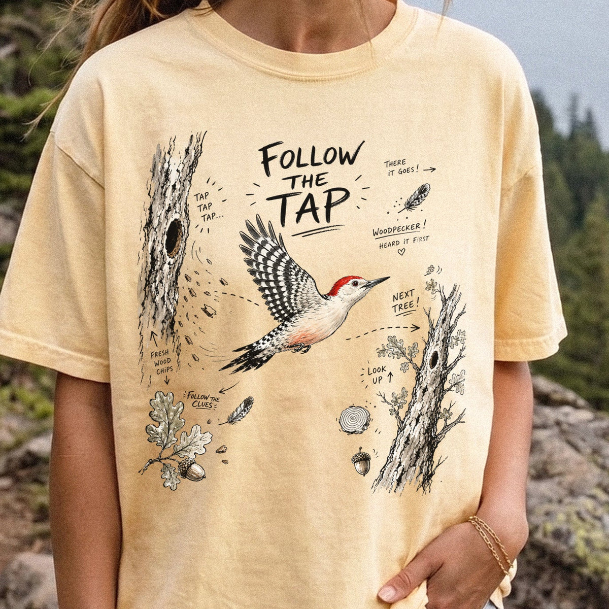 Follow the Tap Comfort Colors Shirt – Red-Bellied Woodpecker design of a Red-Bellied Woodpecker illustrated in flight, surrounded by tree trunks, wood chips, feathers, oak leaves, and acorn details, printed on a Butter T-shirt, worn by a woman.