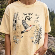 Follow the Tap Comfort Colors Shirt – Red-Bellied Woodpecker design of a Red-Bellied Woodpecker illustrated in flight, surrounded by tree trunks, wood chips, feathers, oak leaves, and acorn details, printed on a Butter T-shirt, worn by a woman.