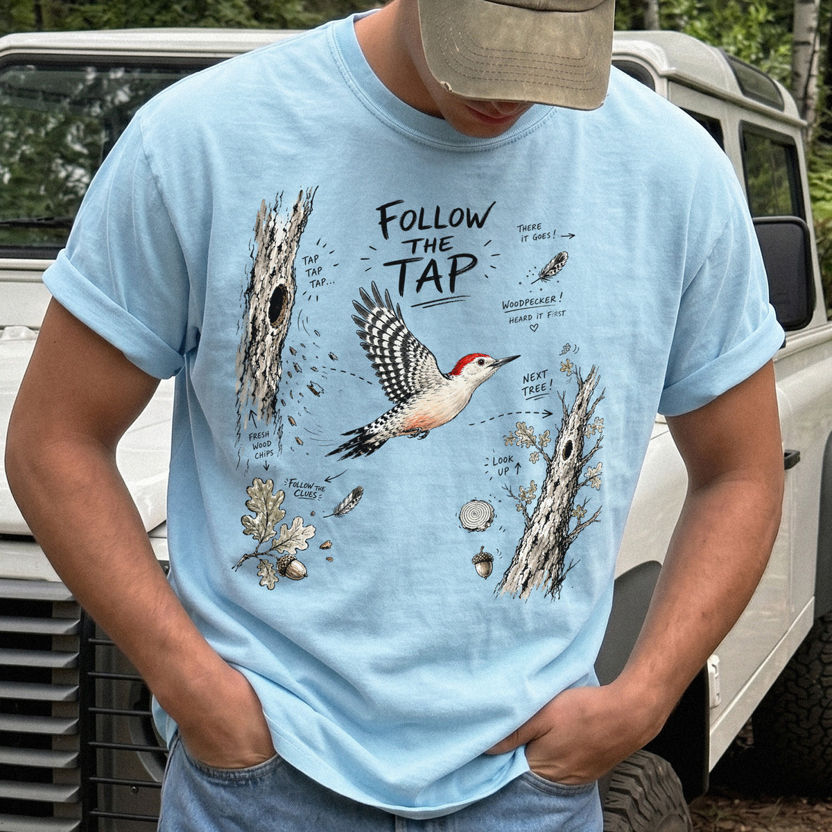 Follow the Tap Comfort Colors Shirt – Red-Bellied Woodpecker design of a Red-Bellied Woodpecker illustrated in flight, surrounded by tree trunks, wood chips, feathers, oak leaves, and acorn details, printed on a Chambray T-shirt.