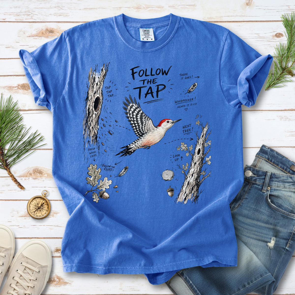 Follow the Tap Comfort Colors Shirt – Red-Bellied Woodpecker design of a Red-Bellied Woodpecker illustrated in flight, surrounded by tree trunks, wood chips, feathers, oak leaves, and acorn details, printed on a Flo Blue T-shirt.