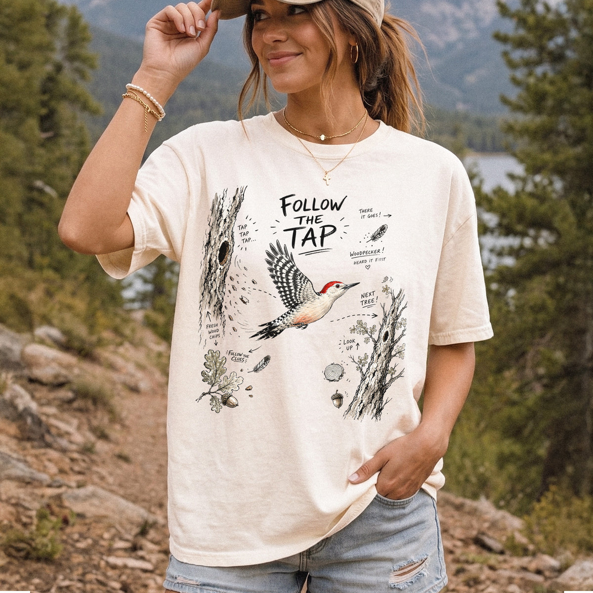 Follow the Tap Comfort Colors Shirt – Red-Bellied Woodpecker design of a Red-Bellied Woodpecker illustrated in flight, surrounded by tree trunks, wood chips, feathers, oak leaves, and acorn details, printed on an Ivory T-shirt, worn by a woman.
