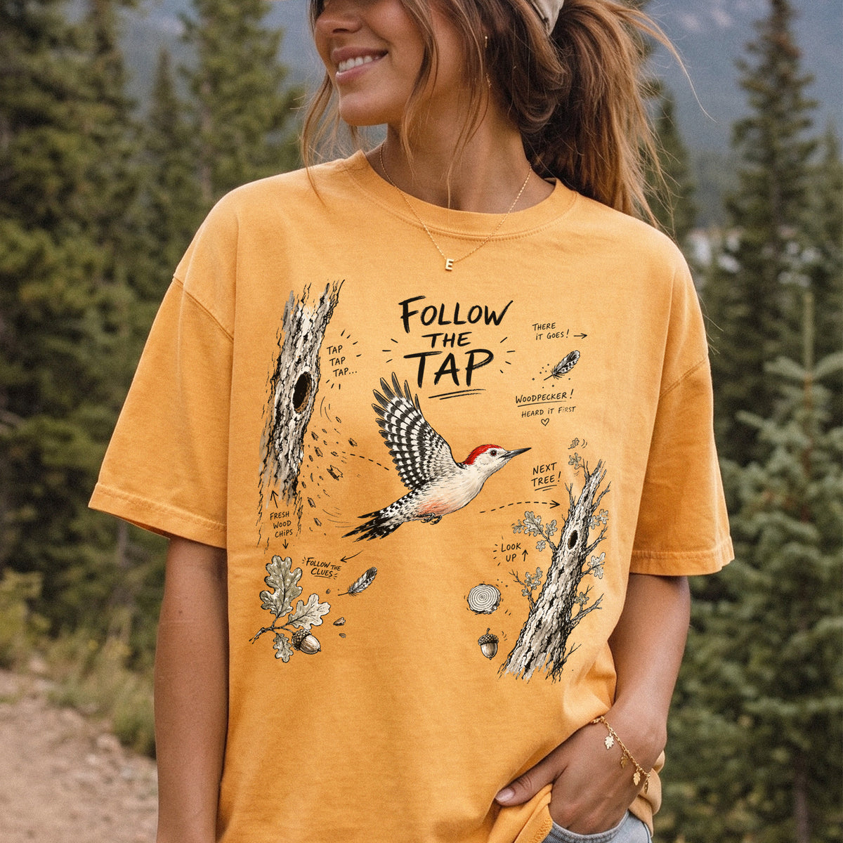 Follow the Tap Comfort Colors Shirt – Red-Bellied Woodpecker design of a Red-Bellied Woodpecker illustrated in flight, surrounded by tree trunks, wood chips, feathers, oak leaves, and acorn details, printed on a Mustard T-shirt, worn by a woman.