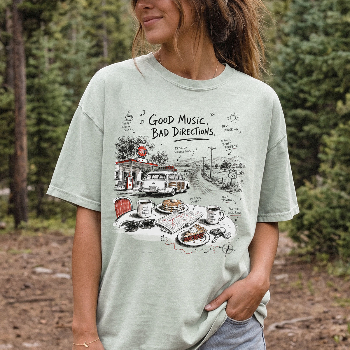 Woman wearing a Bay Comfort Colors T-shirt printed with a vintage roadside scene with a classic car, old gas station, and a winding back road
