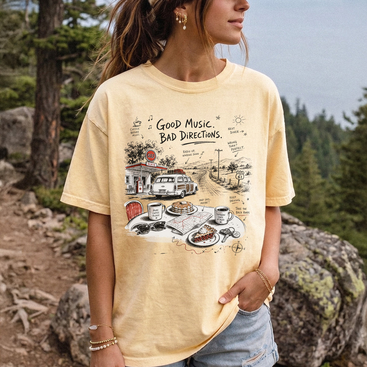 Woman wearing a Butter Comfort Colors T-shirt printed with a vintage roadside scene with a classic car, old gas station, and a winding back road