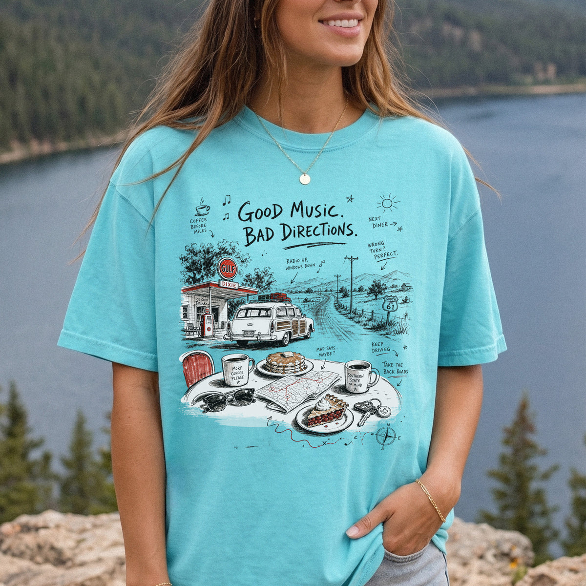 Woman wearing a Chalky Mint Comfort Colors T-shirt printed with a vintage roadside scene with a classic car, old gas station, and a winding back road