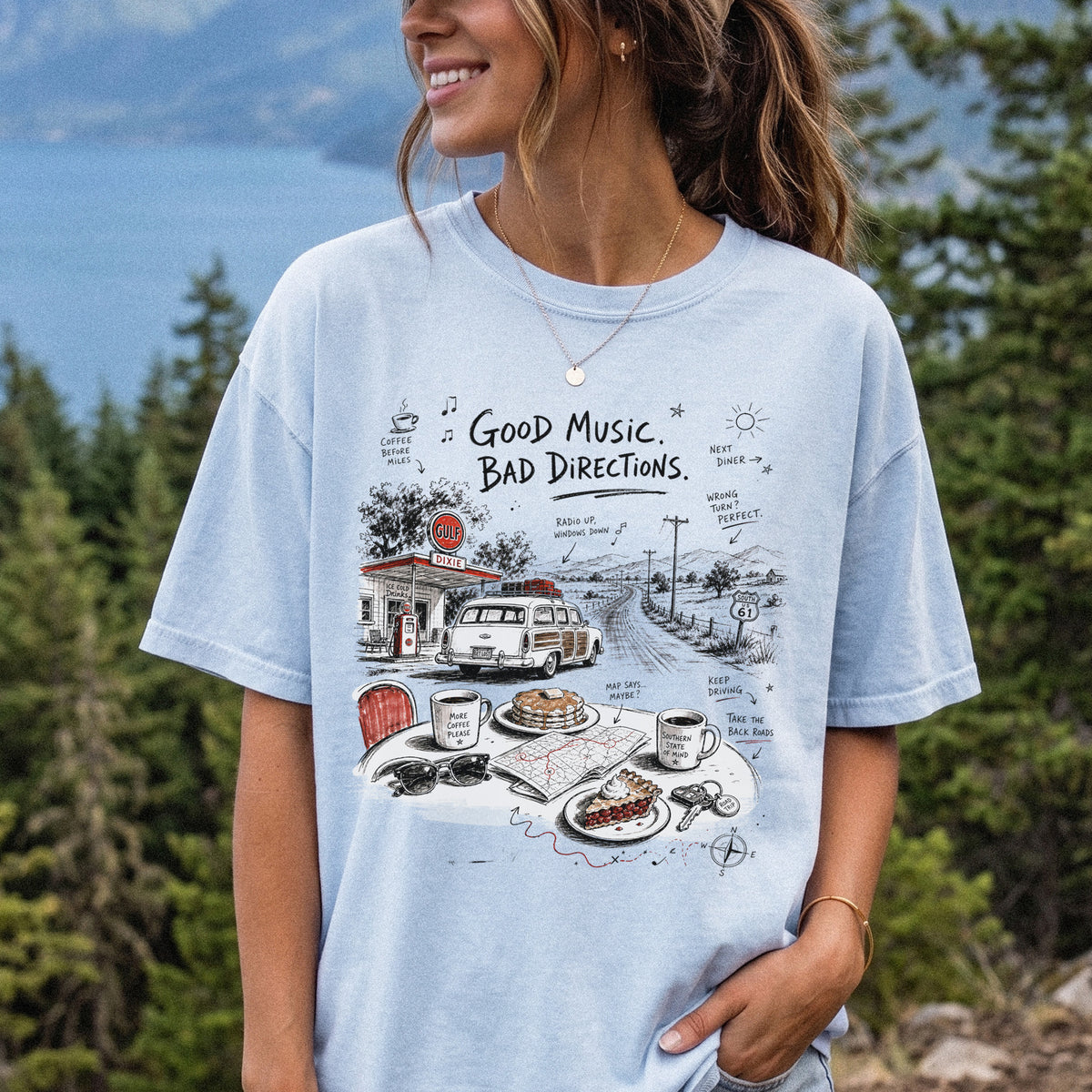 Woman wearing a Chambray Comfort Colors T-shirt printed with a vintage roadside scene with a classic car, old gas station, and a winding back road