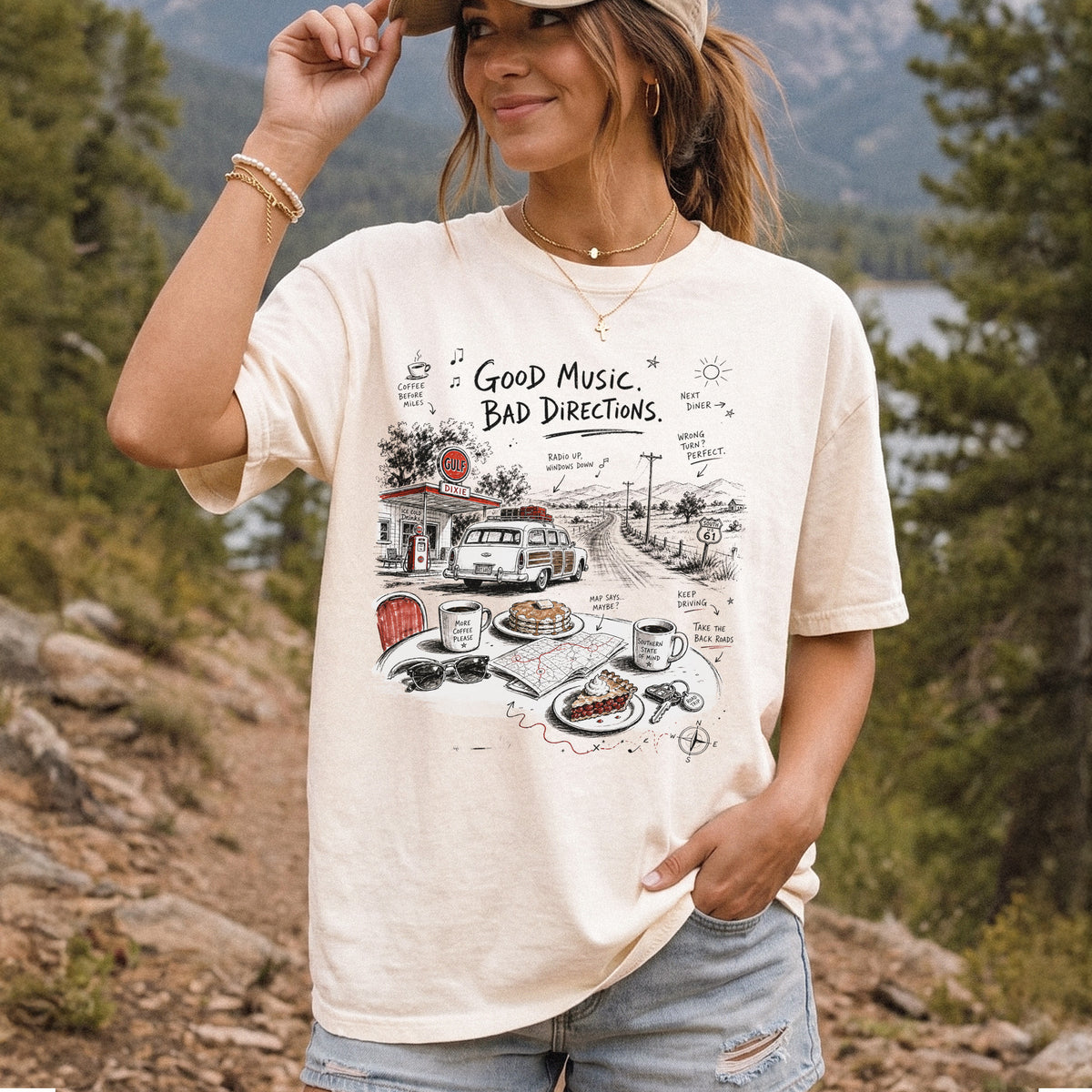 Woman wearing an Ivory Comfort Colors T-shirt printed with a vintage roadside scene with a classic car, old gas station, and a winding back road