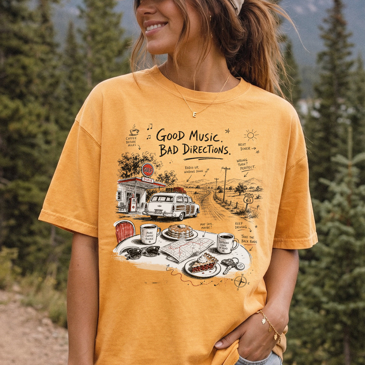 Woman wearing a Mustard Comfort Colors T-shirt printed with a vintage roadside scene with a classic car, old gas station, and a winding back road