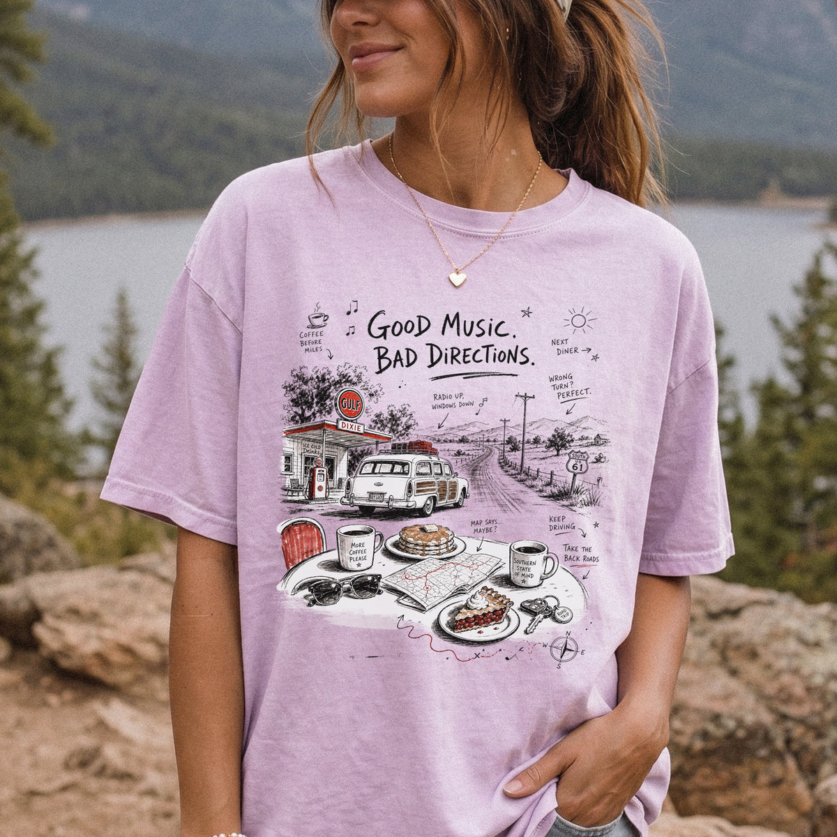 Woman wearing an Orchid Comfort Colors T-shirt printed with a vintage roadside scene with a classic car, old gas station, and a winding back road