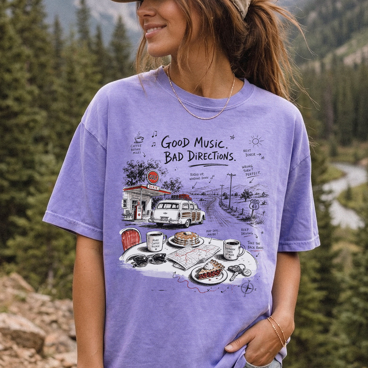 Woman wearing a Violet Comfort Colors T-shirt printed with a vintage roadside scene with a classic car, old gas station, and a winding back road