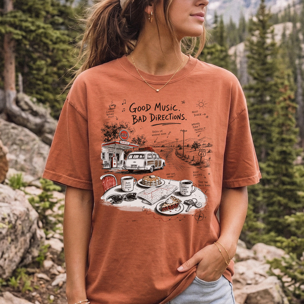 Woman wearing a Yam Comfort Colors T-shirt printed with a vintage roadside scene with a classic car, old gas station, and a winding back road