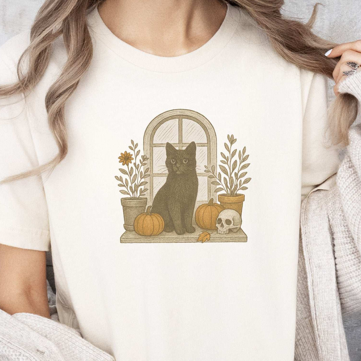 Woman wearing a Natural T-shirt printed with a black cat perched in a window among pumpkins, plants, and a skull