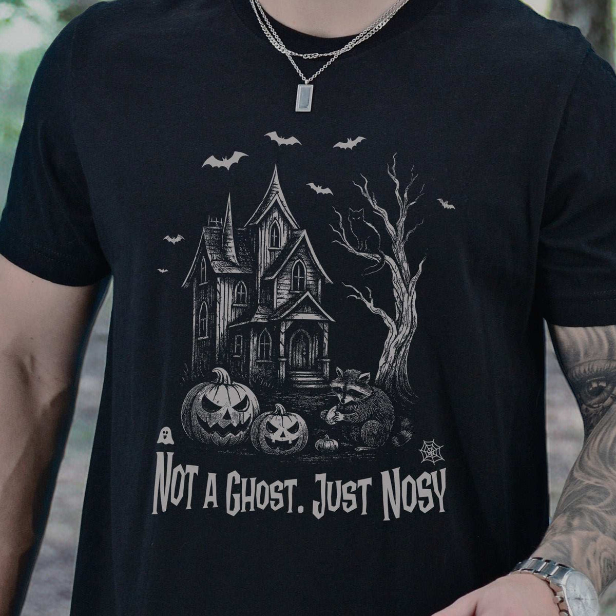 Man wearing a Black T-shirt printed with a raccoon outside a haunted house with pumpkins, bats, and a cat, gothic illustration