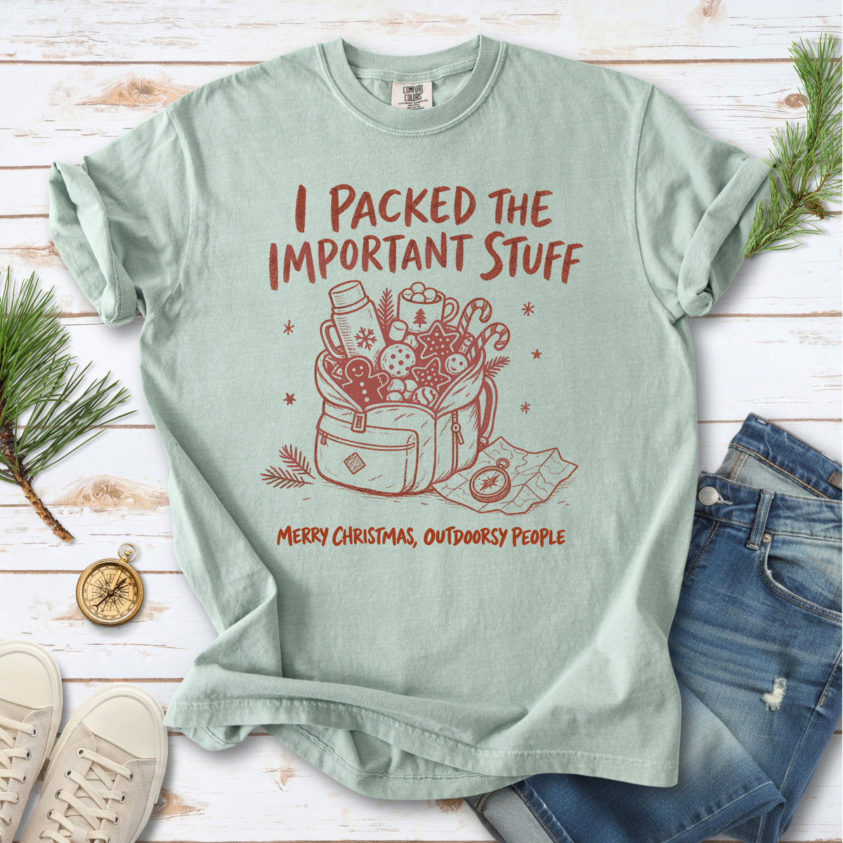 An overstuffed backpack with a thermos, mug, compass, trail map, and festive treats, doodle style design on a Bay T-shirt