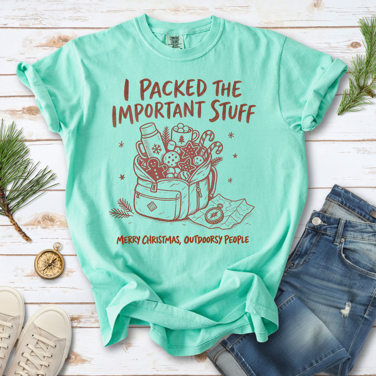 An overstuffed backpack with a thermos, mug, compass, trail map, and festive treats, doodle style design on an Island Reef T-shirt