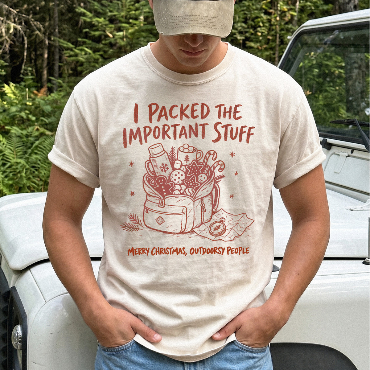 Man wearing an Ivory T-shirt printed with an overstuffed backpack with a thermos, mug, compass, trail map, and festive treats, doodle style