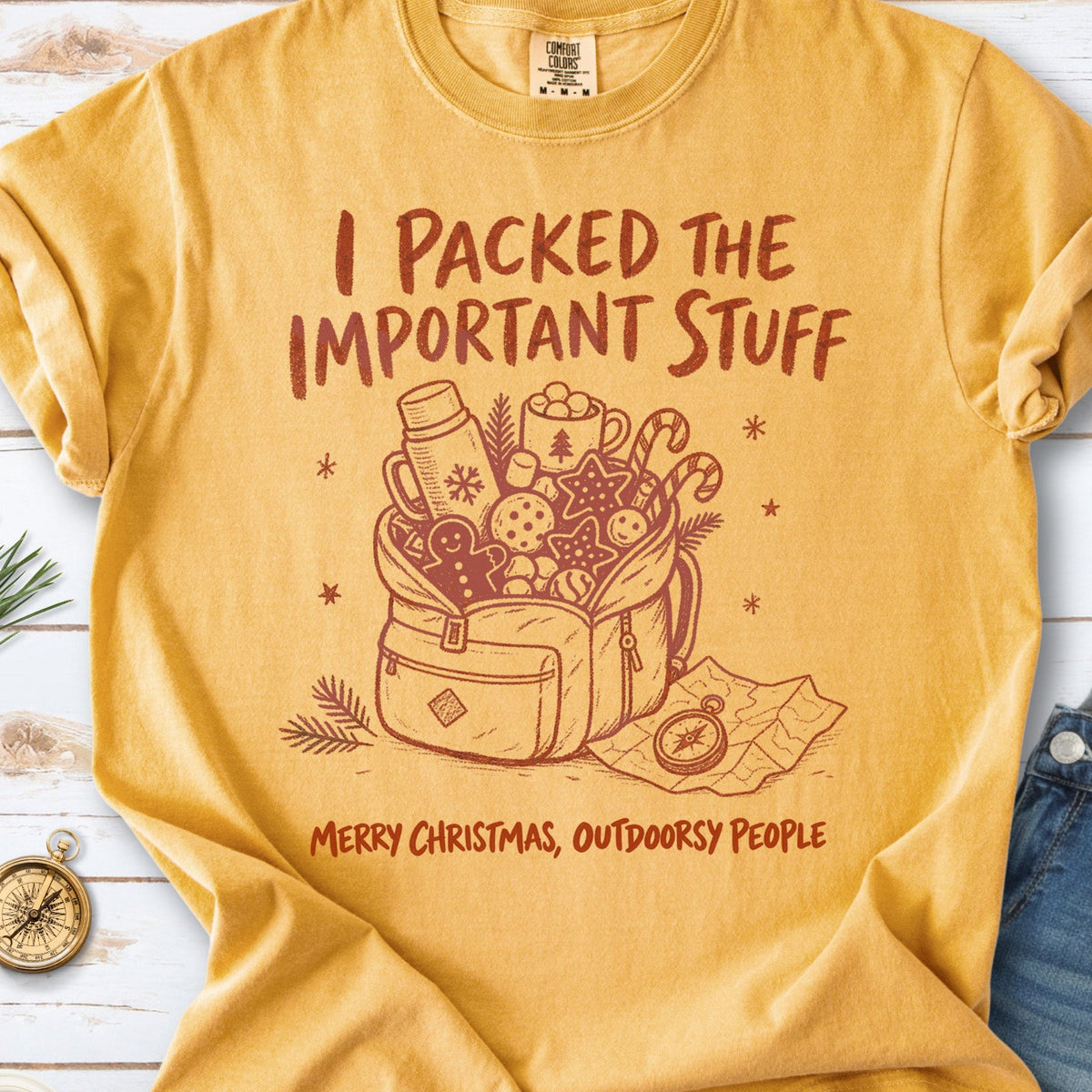 An overstuffed backpack with a thermos, mug, compass, trail map, and festive treats, doodle style design on a Mustard T-shirt