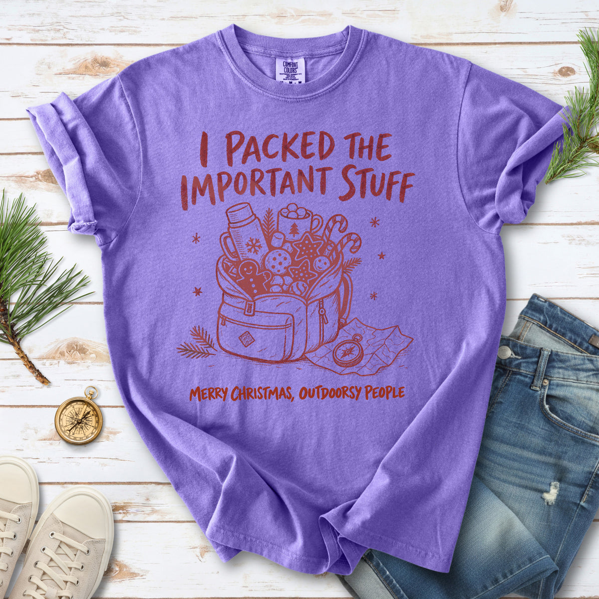 An overstuffed backpack with a thermos, mug, compass, trail map, and festive treats, doodle style design on a Violet T-shirt