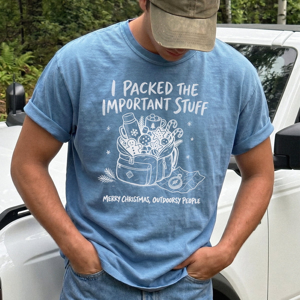 Man wearing a Blue Jean T-shirt printed with an overstuffed backpack with a thermos, mug, compass, trail map, and festive treats, doodle style