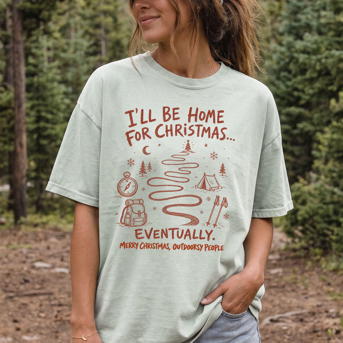 Woman wearing a Bay T-shirt printed with a winding trail with a tent, compass, backpack, trekking poles, snowflakes, and pine trees, doodle style
