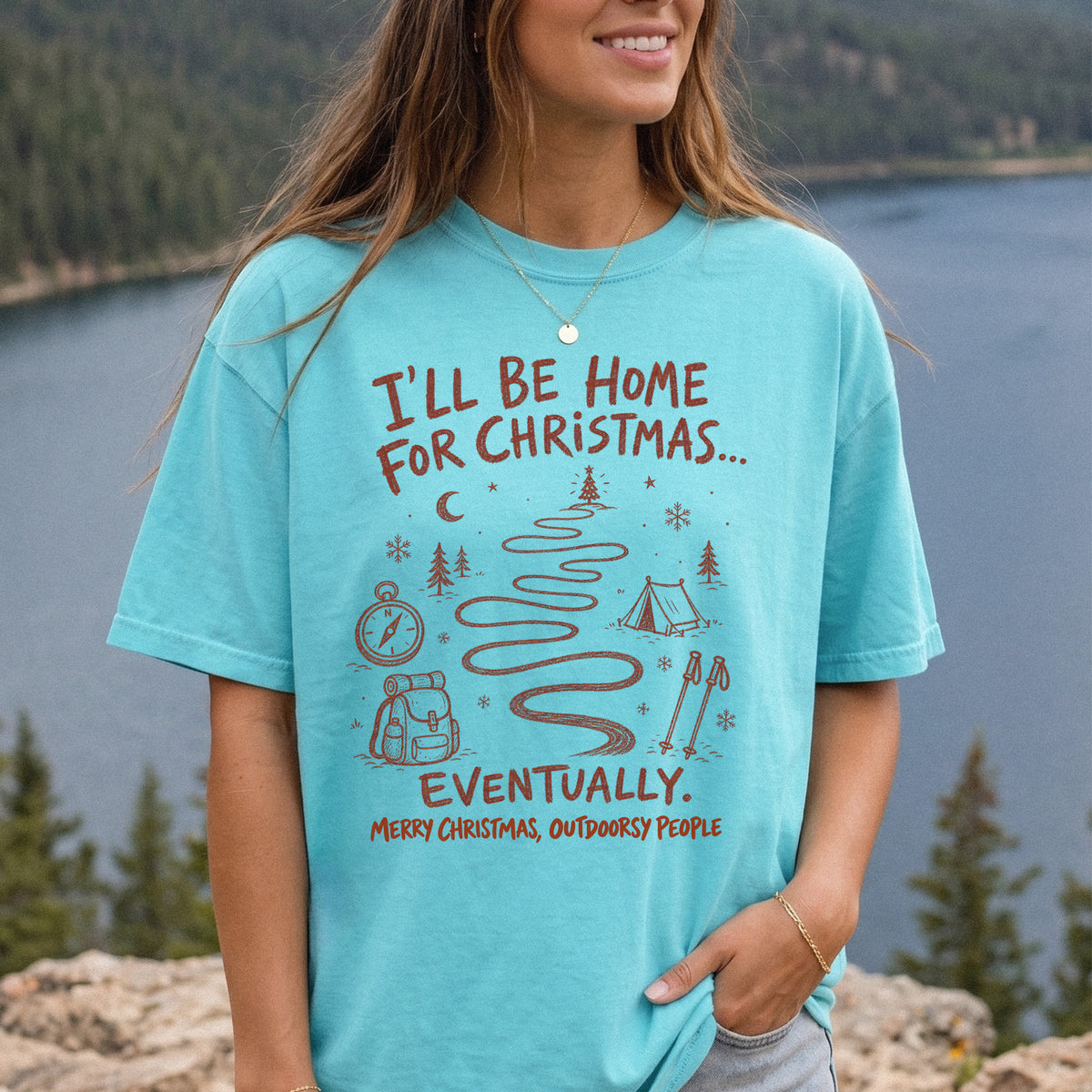 Woman wearing a Chalky Mint T-shirt printed with a winding trail with a tent, compass, backpack, trekking poles, snowflakes, and pine trees, doodle style