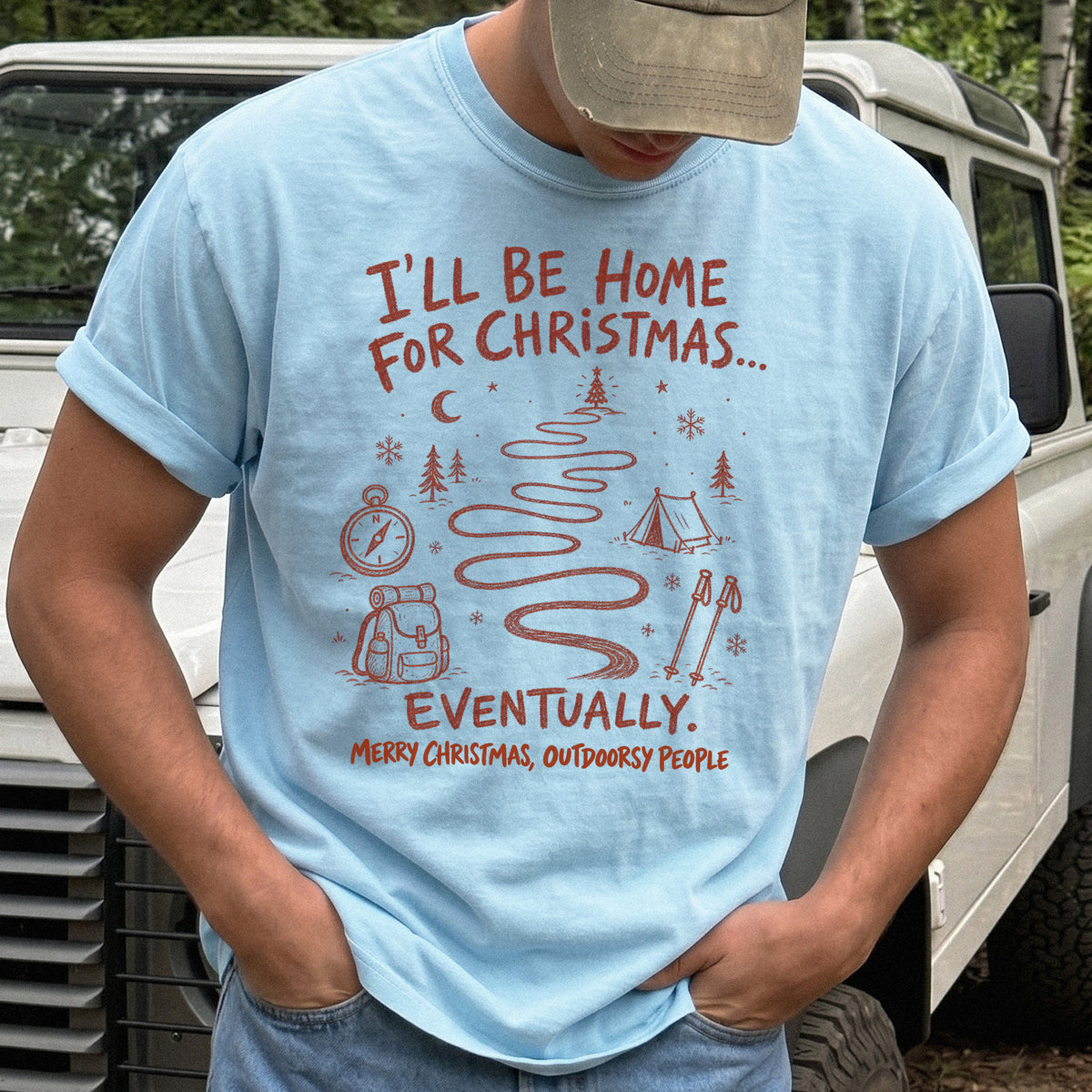 Man wearing a Chambray T-shirt printed with a winding trail with a tent, compass, backpack, trekking poles, snowflakes, and pine trees, doodle style