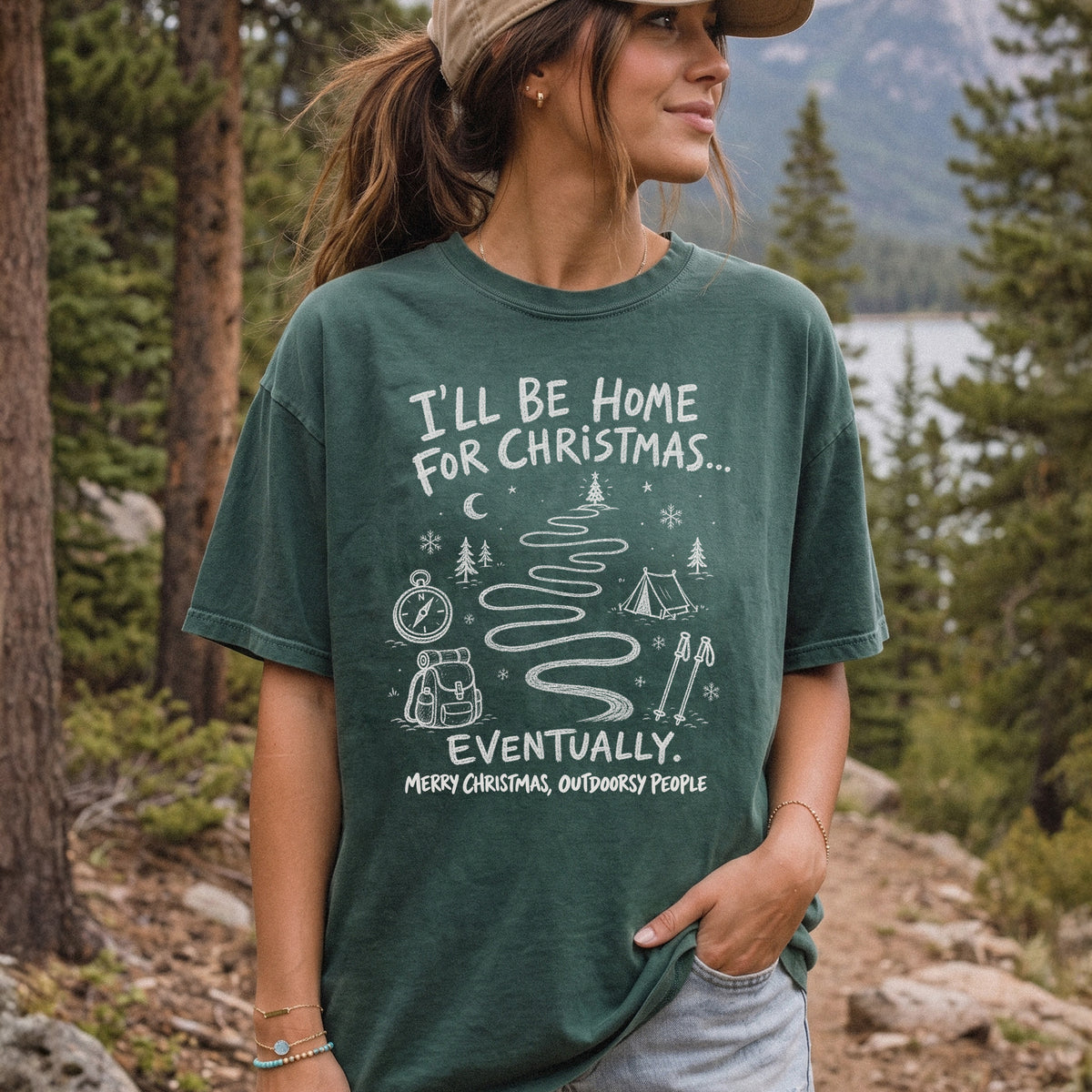 Woman wearing a Blue Spruce T-shirt printed with a winding trail with a tent, compass, backpack, trekking poles, snowflakes, and pine trees, doodle style