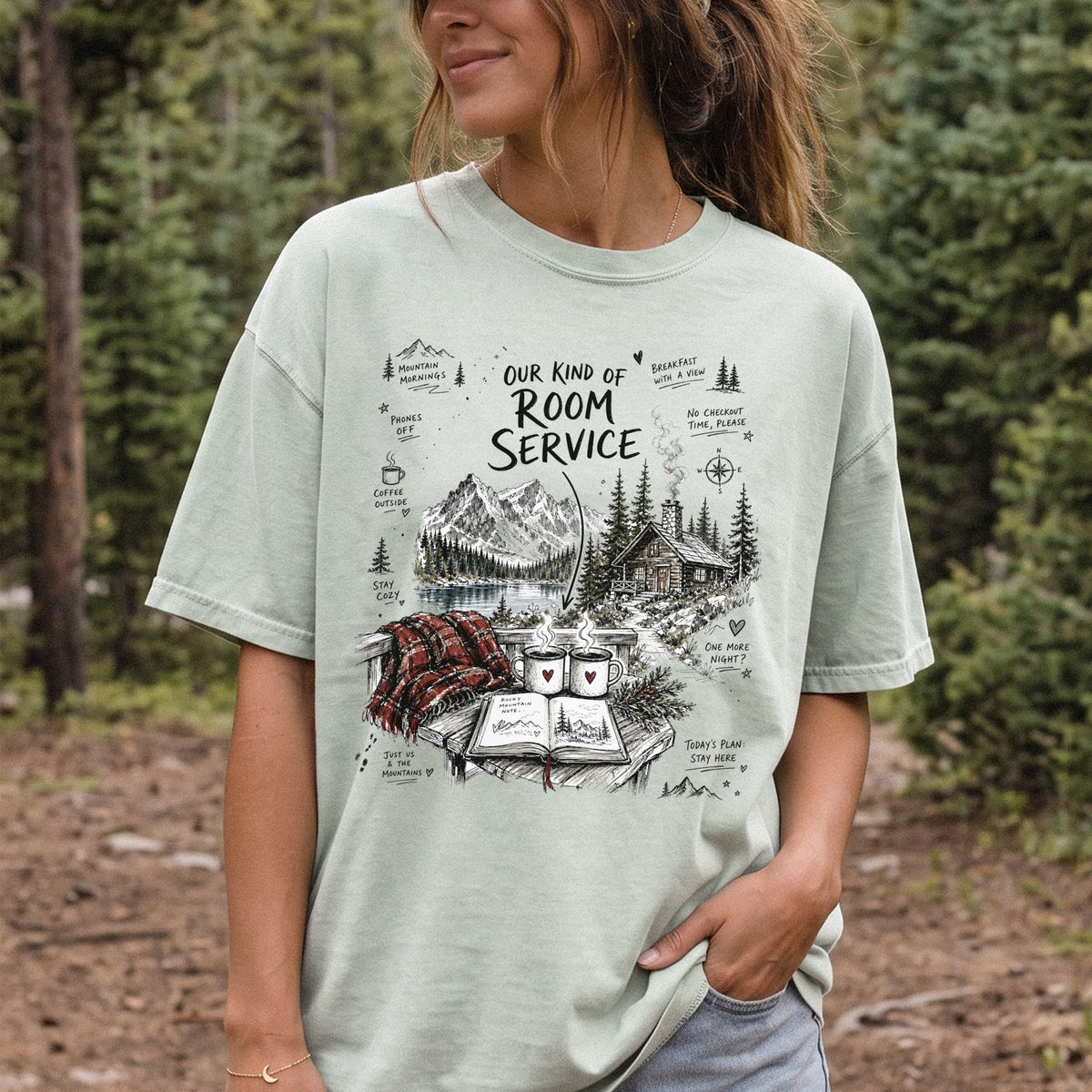 Woman wearing a Bay Comfort Colors T-shirt printed with a cozy lakeside cabin scene with Rocky Mountain peaks, coffee mugs, and a journal