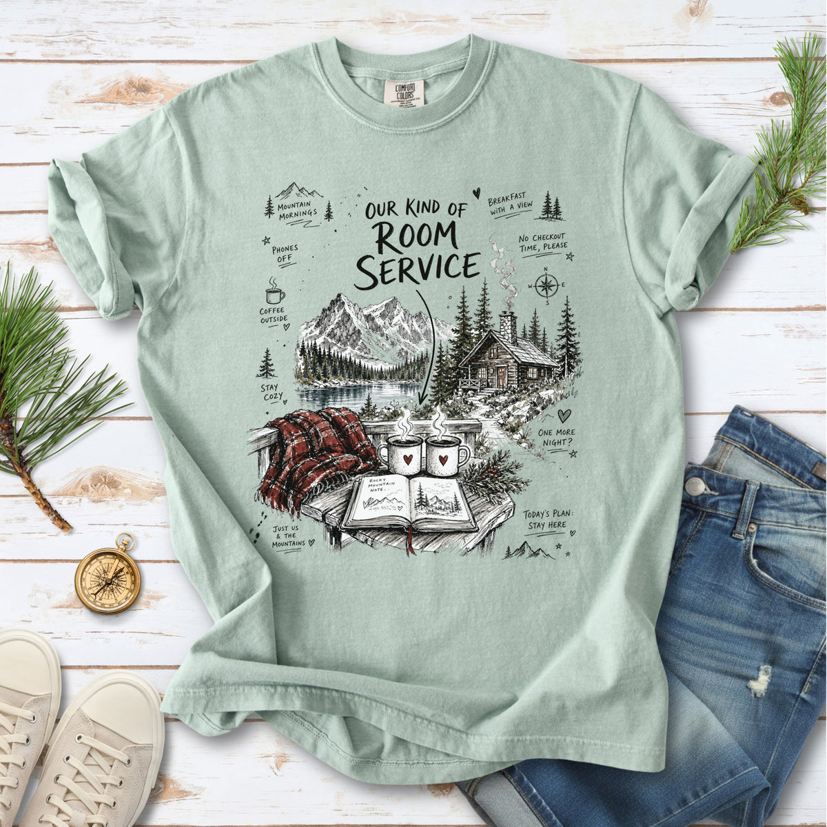 A cozy lakeside cabin scene with Rocky Mountain peaks, coffee mugs, and a journal design on a Bay Comfort Colors T-shirt