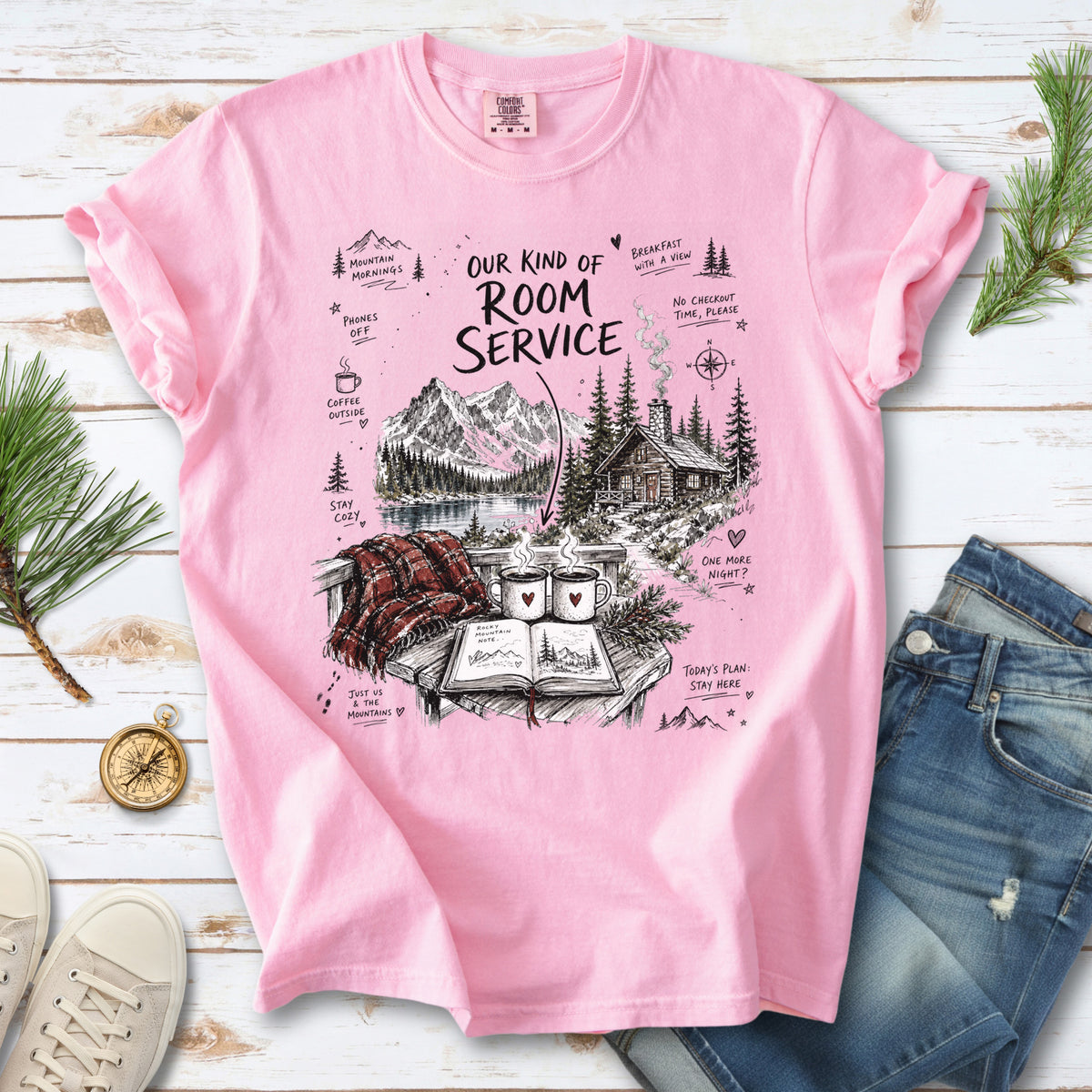 A cozy lakeside cabin scene with Rocky Mountain peaks, coffee mugs, and a journal design on a Blossom Comfort Colors T-shirt