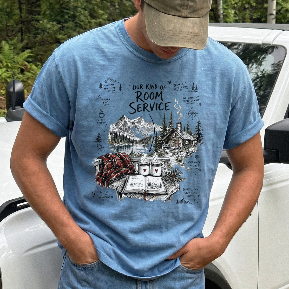 Man wearing a Blue Jean Comfort Colors T-shirt with a cozy lakeside cabin scene with Rocky Mountain peaks, coffee mugs, and a journal