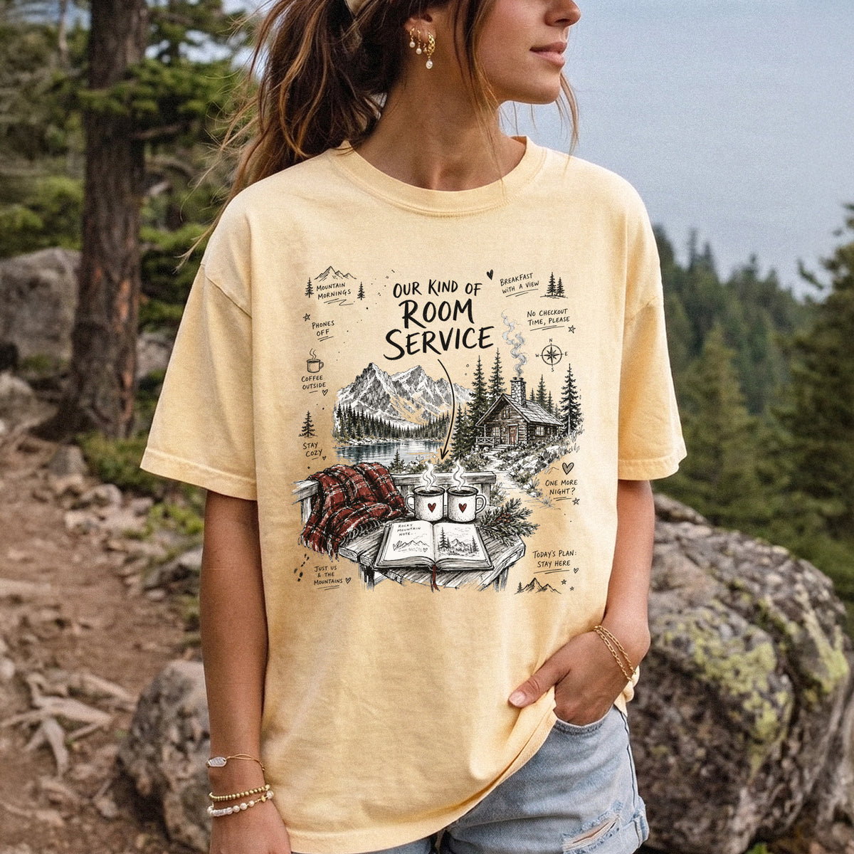 Woman wearing a Butter Comfort Colors T-shirt printed with a cozy lakeside cabin scene with Rocky Mountain peaks, coffee mugs, and a journal