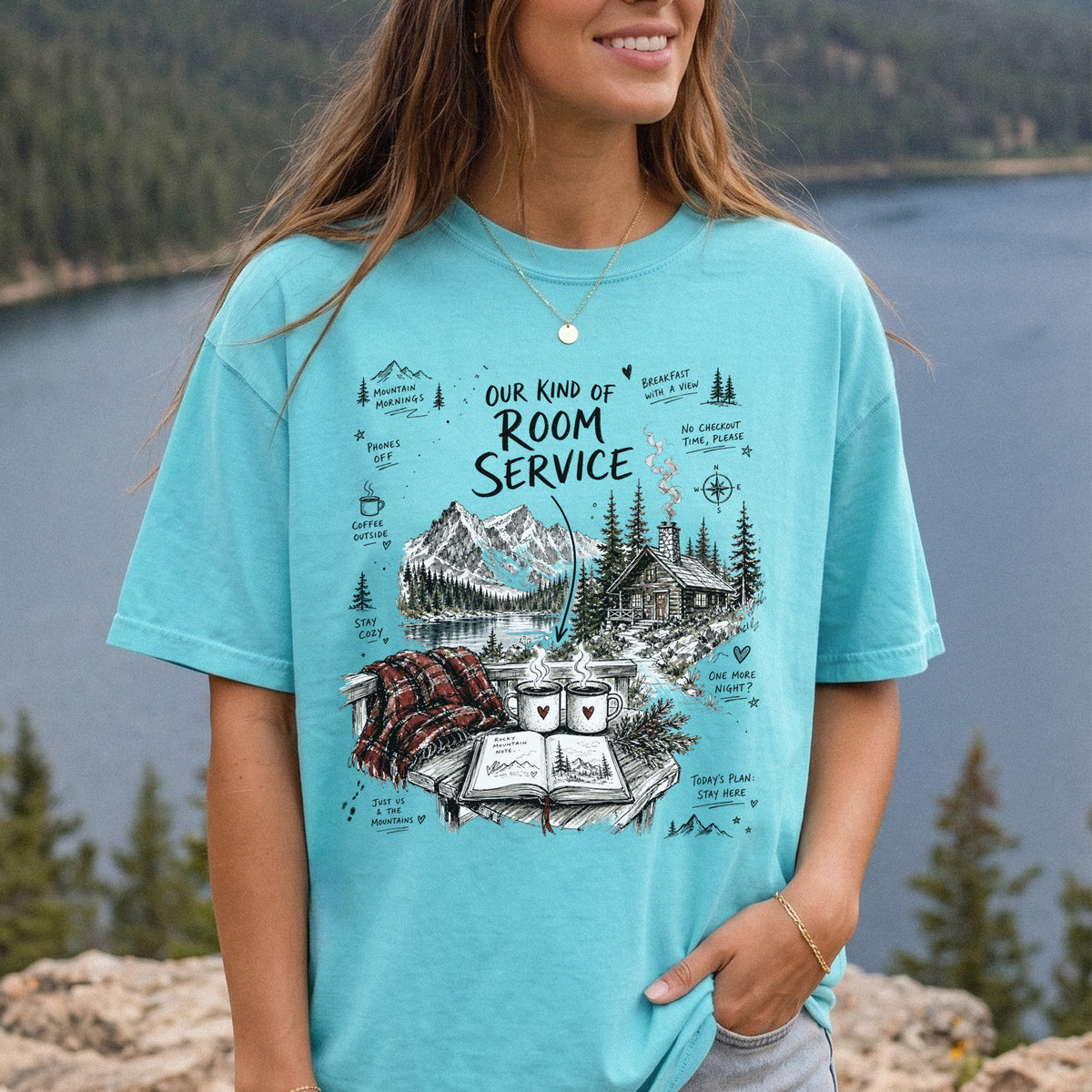 Woman wearing a Chalky Mint Comfort Colors T-shirt printed with a cozy lakeside cabin scene with Rocky Mountain peaks, coffee mugs, and a journal