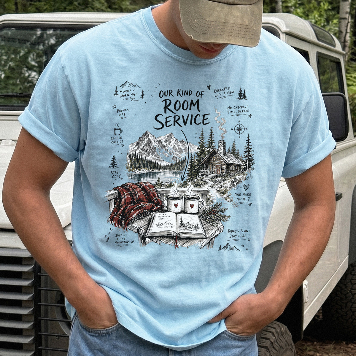 Man wearing a Chambray Comfort Colors T-shirt with a cozy lakeside cabin scene with Rocky Mountain peaks, coffee mugs, and a journal
