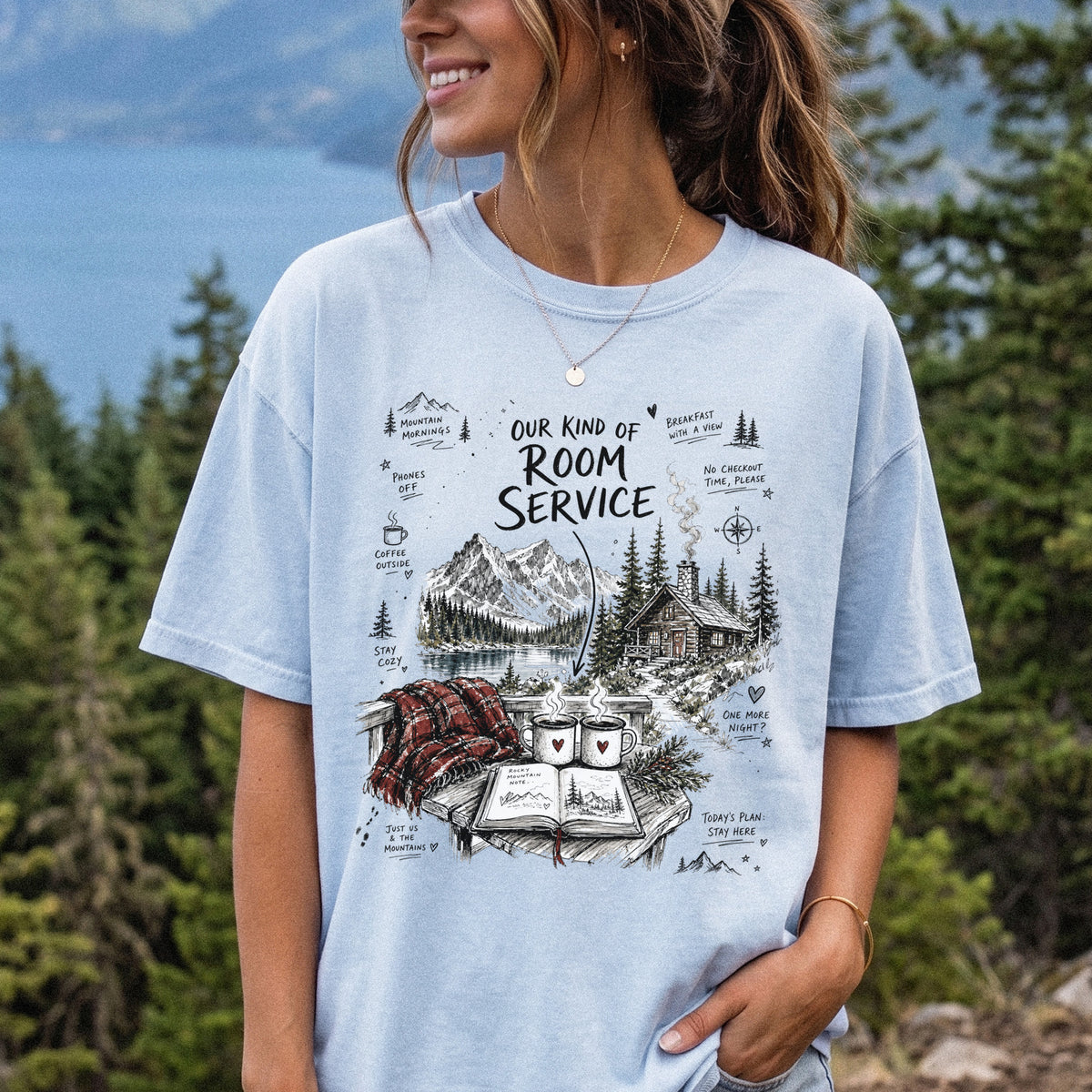 Woman wearing a Chambray Comfort Colors T-shirt printed with a cozy lakeside cabin scene with Rocky Mountain peaks, coffee mugs, and a journal