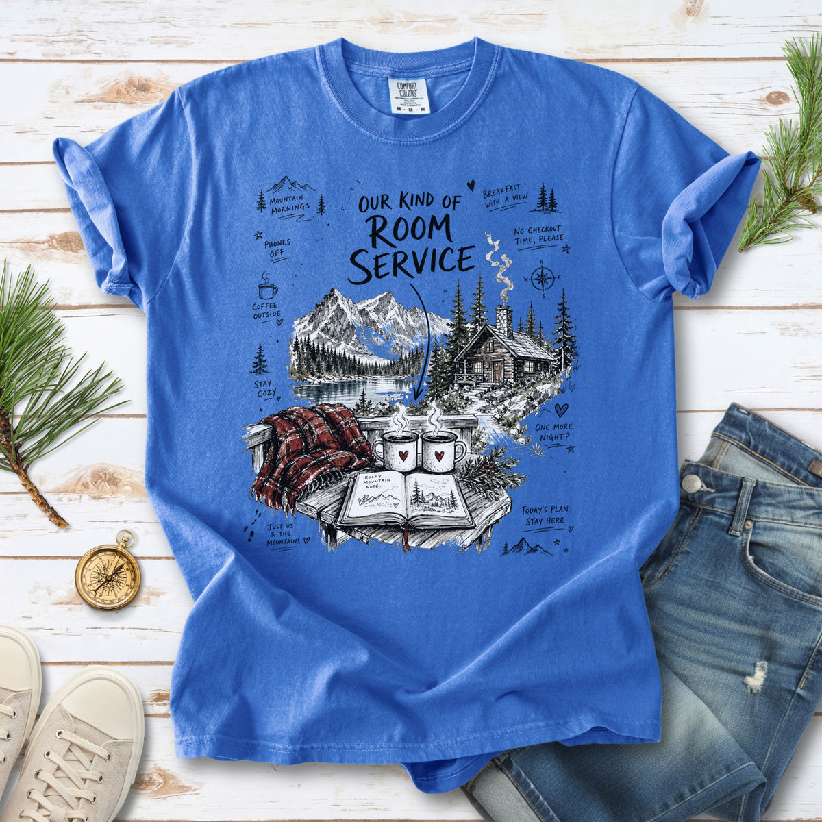 A cozy lakeside cabin scene with Rocky Mountain peaks, coffee mugs, and a journal design on a Flo Blue Comfort Colors T-shirt