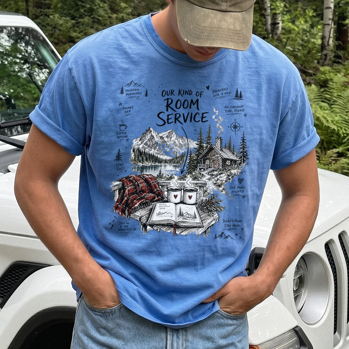 Man wearing a Flo Blue Comfort Colors T-shirt with a cozy lakeside cabin scene with Rocky Mountain peaks, coffee mugs, and a journal