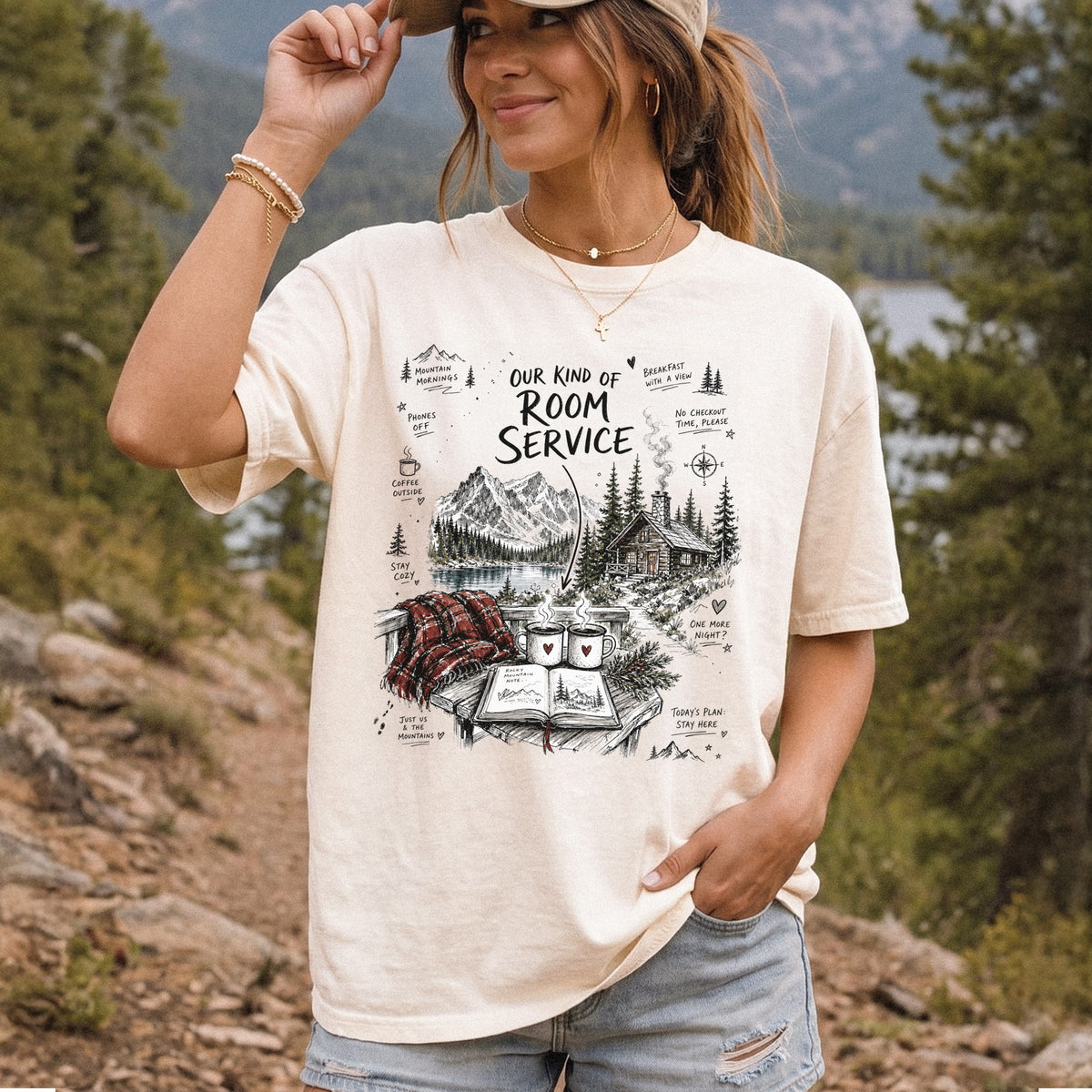 Woman wearing an Ivory Comfort Colors T-shirt printed with a cozy lakeside cabin scene with Rocky Mountain peaks, coffee mugs, and a journal