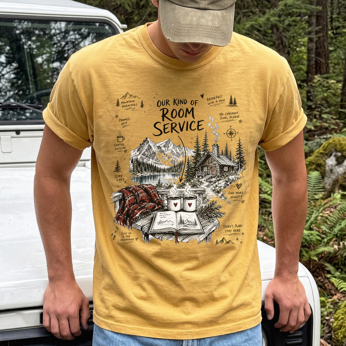Man wearing a Mustard Comfort Colors T-shirt with a cozy lakeside cabin scene with Rocky Mountain peaks, coffee mugs, and a journal